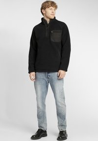 Czarny polarowy sweter z półzamkiem, z zamykaną na zamek kieszenią na klatce piersiowej i teksturowanym materiałem. Noszony z jasnoniebieskimi dżinsami i czarnymi butami.