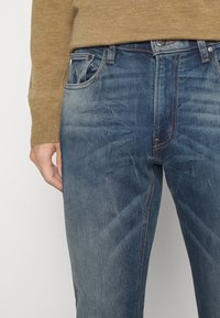 Modré džínové kalhoty s vybledlým povrchem, které mají pět kapes, kontrastní švy a zipový létal. Materiál se zdá být měkký s střední váhou.