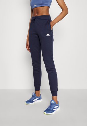 Persoon draagt marineblauwe Adidas joggingbroek, blauwe Adidas sneakers en een blauwe sportbeha, staand tegen een effen lichte achtergrond.