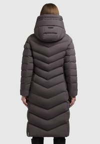 Frau, die einen langen, dunkelgrauen gepolsterten Wintermantel mit einer großen Kapuze trägt, steht mit dem Rücken zu einem hellen, schlichten Hintergrund.