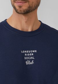 Donkerblauwe sweatshirt met een ronde halslijn, voorzien van witte geborduurde tekst: "LONESOME RIDER SOCIAL Club." Katoenen stof met een zachte textuur.
