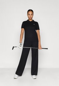 Polo negro de punto trenzado con manga corta y cuello, combinado con pantalones anchos a juego. El modelo sostiene un palo de golf, llevando un guante y zapatillas.