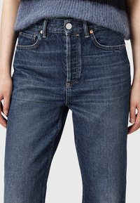 Jeans in denim blu scuro con design a gamba dritta, vita alta e tasche frontali. Dettagliati con cuciture a contrasto e un bottone in metallo.