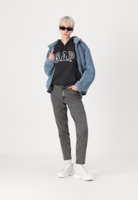 Jachetă din fleece albastru deschis peste o hanorac negru cu logo-ul "GAP", blugi gri și adidași albi cu talpă groasă. Accesoriile includ ochelari de soare negri.