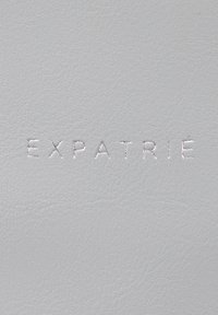 Superficie in pelle grigia con testo in rilievo "EXPATRIE". La texture è liscia con un sottile motivo a grana, creando un aspetto elegante.