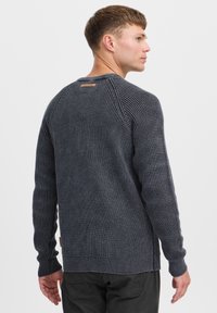 Pull en maille marine avec un motif texturé, des poignets et un ourlet côtelés, et une petite étiquette de marque sur le col arrière.