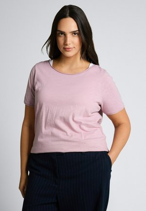 Kobieta o długich ciemnych włosach, ubrana w jasnoróżowy krótki top z krótkim rękawem i ciemne spodnie w cienkie prążki, stoi z jedną ręką w kieszeni na jednolitym tle.