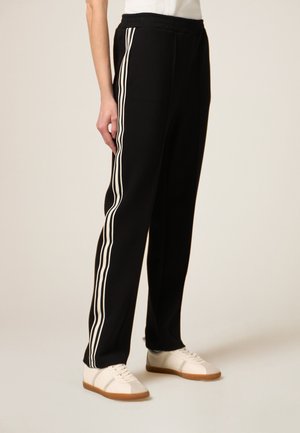 Pantaloni tuta neri con strisce bianche laterali, cintura elastica e vestibilità comoda, abbinati a sneaker bianche con suola in gomma.