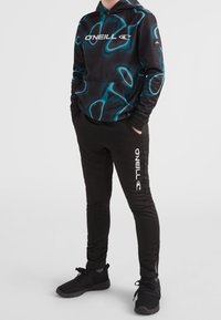 Schwarze Kapuzenjacke mit türkisfarbenem abstraktem Muster, ausgestattet mit einer Fronttasche und dem O'Neill-Logo. Kombiniert mit schwarzen Jogginghosen und schwarzen Sneakers.