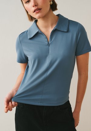 Femme portant un polo bleu à manches courtes avec un col zippé, tenant l'ourlet, debout devant un fond neutre.