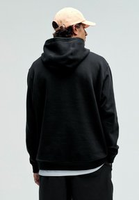Zwarte hoodie met een grote capuchon en lange mouwen, gemaakt van een gestructureerde stof. Draagt een roze pet. Eenvoudig ontwerp, geen zichtbare logo's.