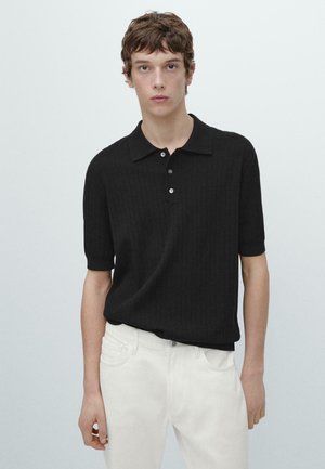 Młody mężczyzna ubrany w czarną, prążkowaną koszulkę polo z krótkim rękawem i białe spodnie, stojący na tle jednolitego, jasnoszarego tła.