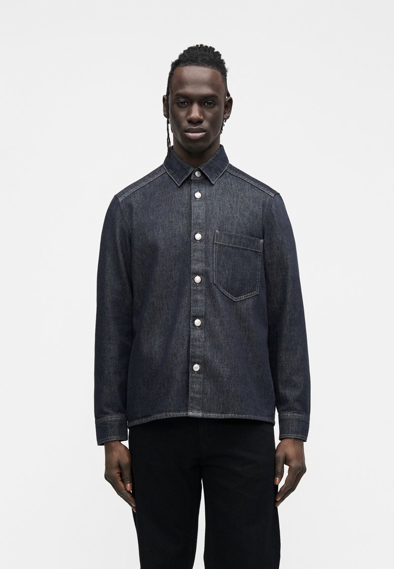 Tmavá džínová košile s knoflíkovou légou vpředu, klasickým límcem, jednou kapsou na prsou a kontrastním šitím. Model nosí tmavé kalhoty.