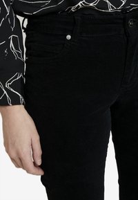 Černé džíny s hladkým povrchem, vybavené předními kapsami, zapínáním na knoflík a detaily v švech. Ruka položená na noze.