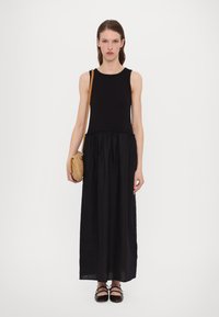 Robe maxi noire sans manches avec un corsage ajusté et une taille froncée. Associée à un sac bandoulière tressé marron et à des chaussures plates noires.