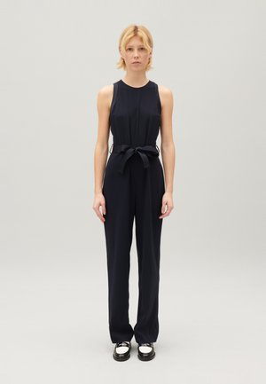 Combinaison sans manches bleu marine avec un corsage ajusté et une taille ceinturée, présentant un pantalon large et un tissu lisse et structuré. Chaussures noires avec des accents métalliques.