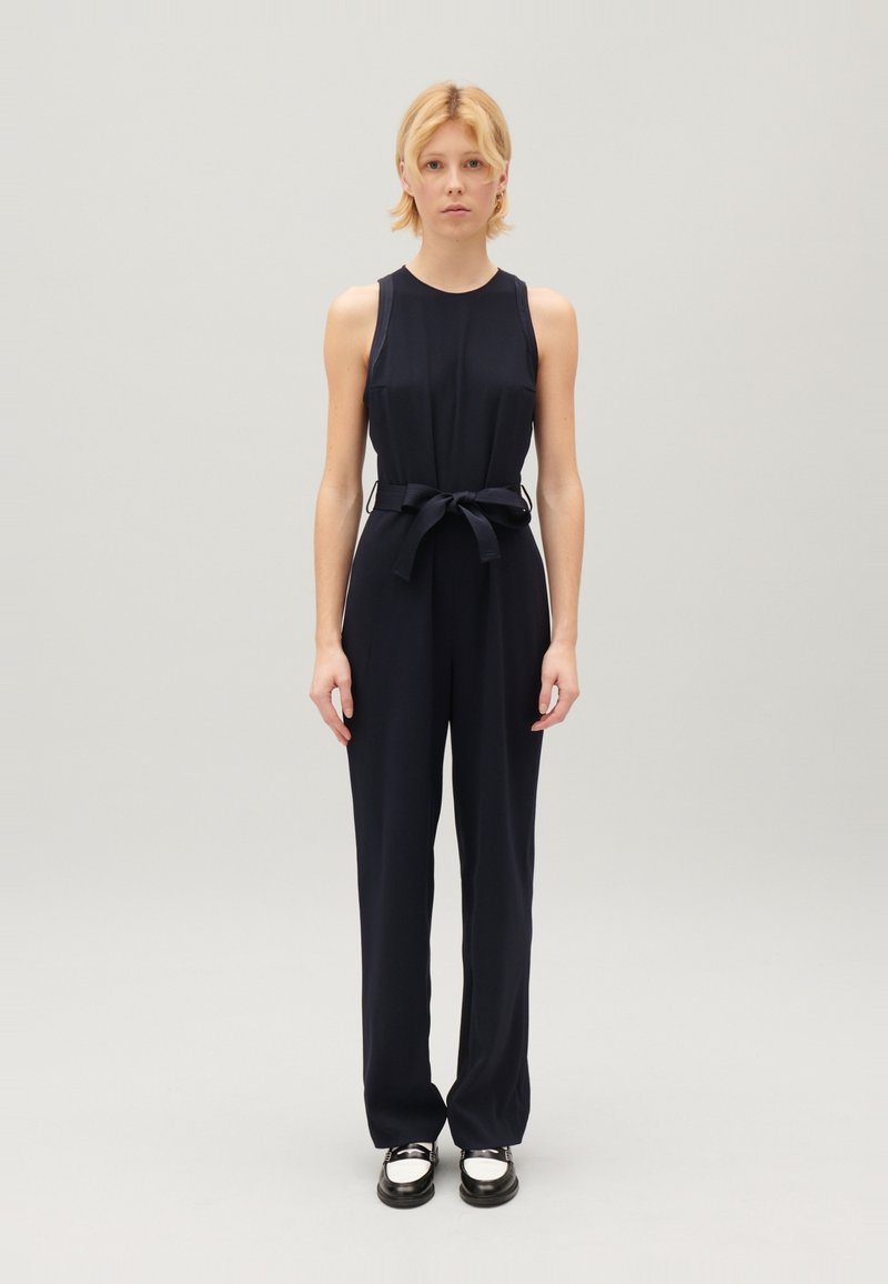 Combinaison sans manches bleu marine avec un corsage ajusté et une taille ceinturée, présentant un pantalon large et un tissu lisse et structuré. Chaussures noires avec des accents métalliques.