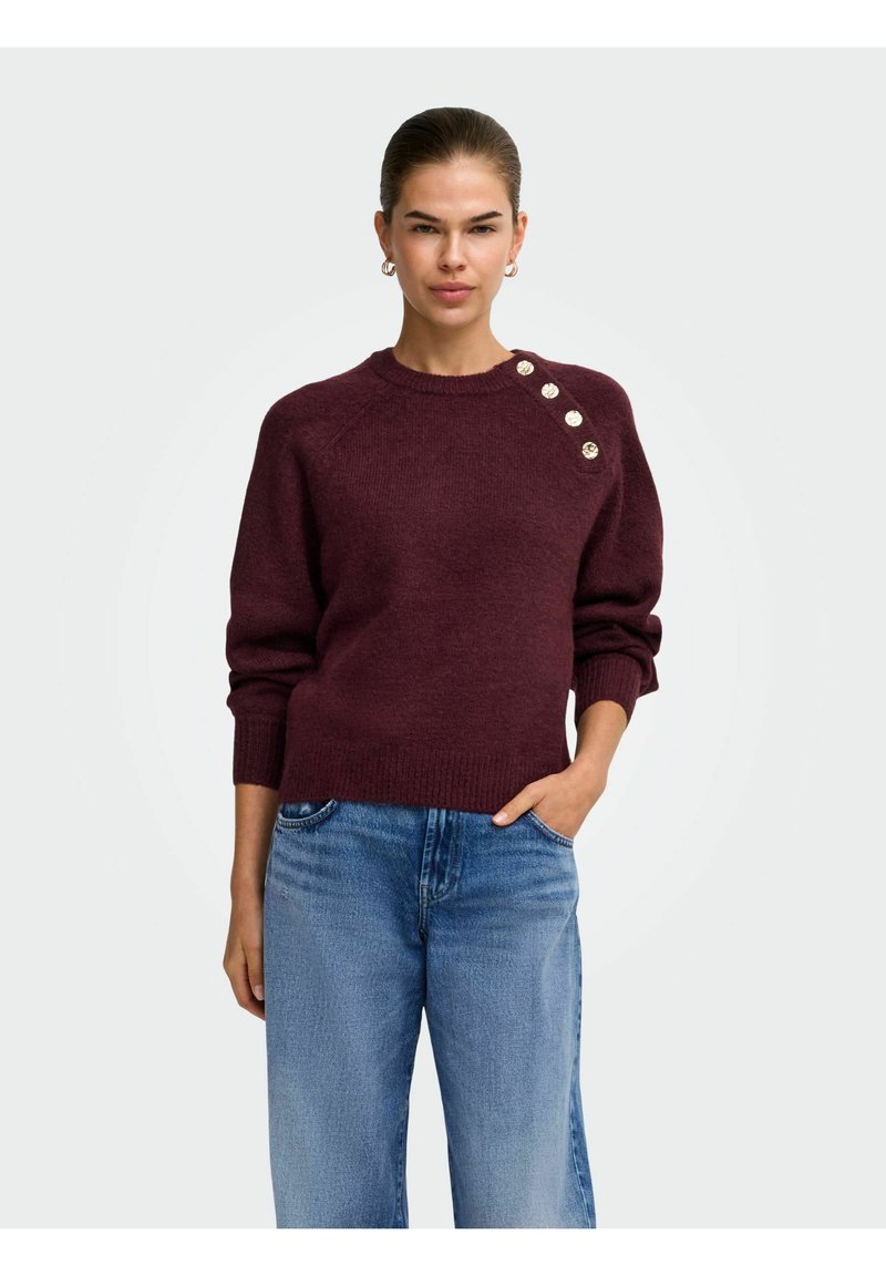 Femme aux cheveux plaqués en arrière portant un pull bordeaux avec des boutons dorés sur l'épaule et un jean bleu, debout avec une main dans la poche.