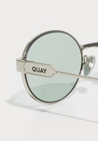 Okrogle sončne naočare s srebrnim okvirjem in svetlo zelenimi lečami ter logotipom "QUAY" na ročaju na belem ozadju.