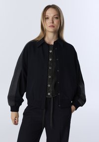 Zwarte bomberjack met een knoopsluiting aan de voorkant, oversized ontwerp en geribbelde manchetten. Heeft contrasterende donkergrijze zijpanelen. Zachte stoftextuur.