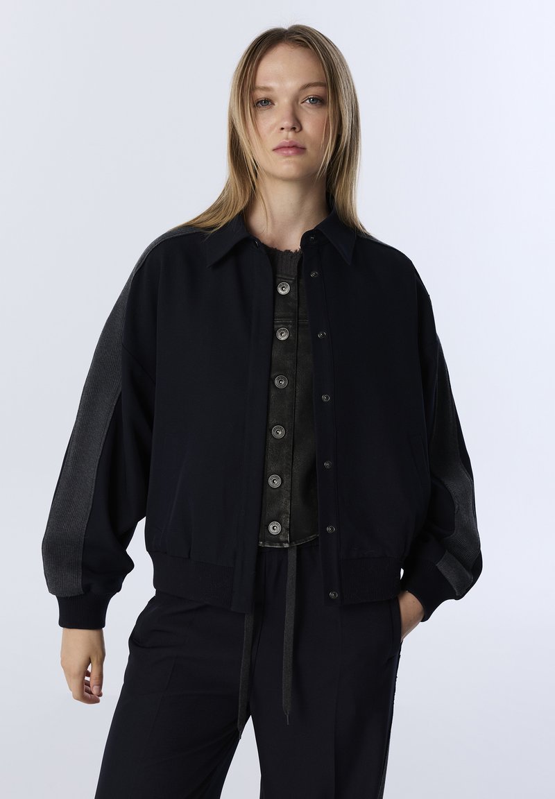 Zwarte bomberjack met een knoopsluiting aan de voorkant, oversized ontwerp en geribbelde manchetten. Heeft contrasterende donkergrijze zijpanelen. Zachte stoftextuur.