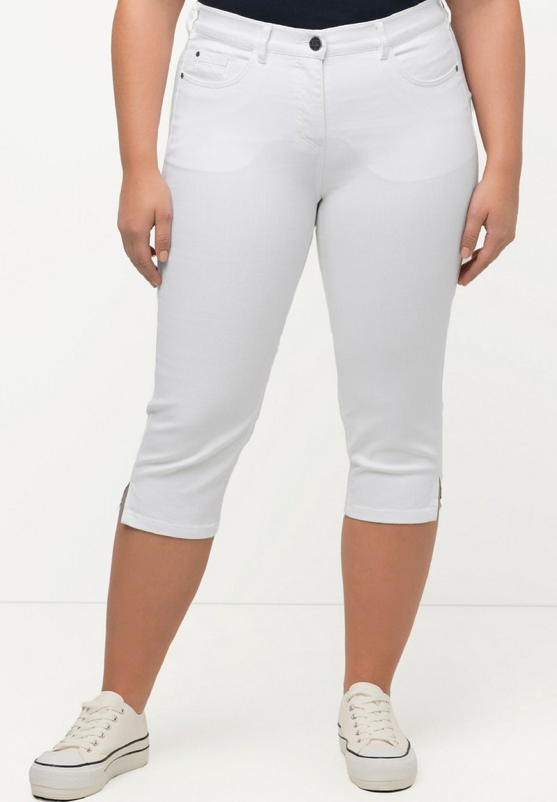 Pantalones capri blancos en denim elástico, con cierre de botón en el frente, cinco bolsillos y aberturas en el dobladillo, combinados con zapatillas de lona blancas.