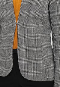 Blazer a quadri grigi e neri parzialmente aperto, che rivela un top testurizzato color senape sotto, ambientato su uno sfondo neutro.