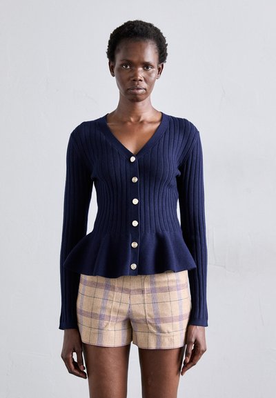 Marineblå ribbet cardigan med V-hals, tætsiddende krop, peplum-nederdel og metalliske knapper. Bærer over ternede shorts. Glat tekstur, lange ærmer.