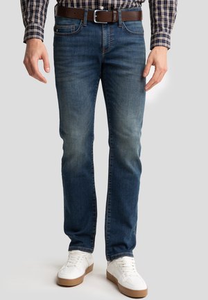 Blue denim jeans with a slim fit, featuring a subtle fade and contrast stitching, worn with a brown belt and white sneakers.