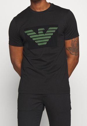 Mann trägt ein schwarzes T-Shirt mit einem grün gestreiften Adler-Logo auf der Brust und ein Schmetterlingstattoo am linken Arm.