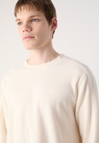 Pulover s krojem crewneck iz mehkega slonokoščenega blaga, z diskretno rebrasto teksturo in majhnim vezenim logotipom na prsih.