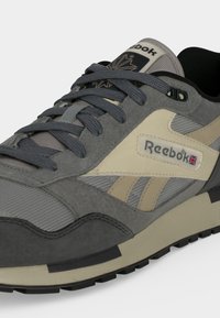 Γκρι και μπεζ αθλητικό παπούτσι Reebok με σκούρα γκρι κορδόνια, υφασμάτινο άνω μέρος με υφή και λογότυπο με μικρή σημαία Union Jack στο πλάι.