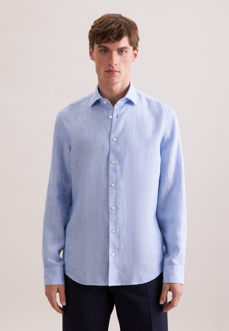 Homme portant une chemise à manches longues bleu clair avec un col classique et un pantalon foncé, debout devant un fond uni.