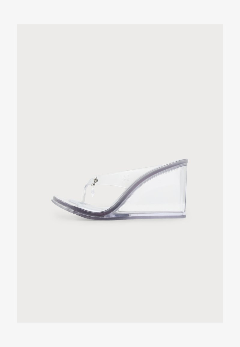 Sandale compensée transparente avec un talon haut transparent et une bride claire en forme de V avec un petit charme décoratif.