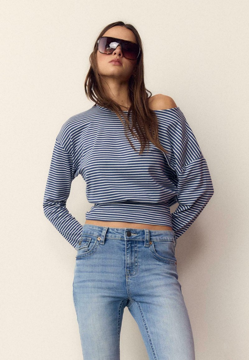 Ung kvinde iført store solbriller, blå- og hvidstribet off-shoulder top og lyseblå højtaljede jeans mod ensfarvet baggrund.