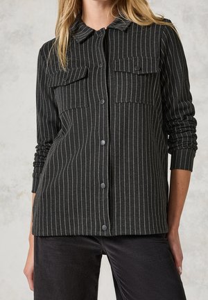 Camisa negra abotonada con rayas verticales blancas, dos bolsillos con solapa en el pecho, combinada con pantalones negros, mangas remangadas en un brazo.