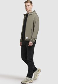 Helle olivgrüne Zip-Jacke mit Kapuze, zwei seitlichen Taschen, elastischen Bündchen, getragen über einem schwarzen Shirt und dunklen Jeans, kombiniert mit hellen Sneakern.