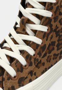 Καφέ καμβάς αθλητικά παπούτσια με σχέδιο λεοπάρ, λευκά κορδόνια, κυκλικές καφέ οπές και υφή από καουτσούκ στη σόλα.