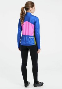 Funkční sportovní bunda v modré, růžové a zelené barvě z polyesteru s vysokým límcem a kapsou na zip na zadní straně. Černé legíny a boty dokreslují celkový vzhled.