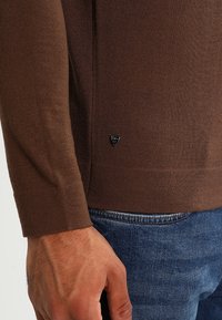 Brun stickad tröja med långa ärmar och en åtsittande design. Försedd med en liten logotyp vid nederkanten. Bärs över blå denimjeans.