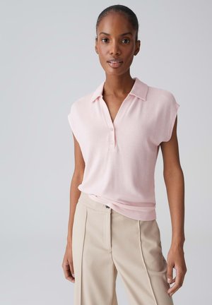 Vrouw met in een knot gebonden haar, gekleed in een lichtroze mouwloze top met kraag en beige broek met hoge taille, tegen een effen achtergrond.