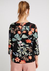 Blouse noire à imprimé floral coloré, manches trois-quarts, avec une fermeture éclair au dos. Tissu lisse avec une coupe décontractée.