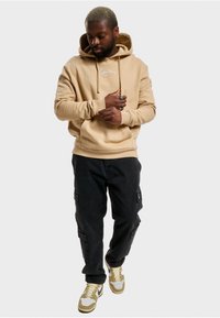 Tan hettegenser med kenguru-lomme og hvit logo, kombinert med svarte cargo-bukser. Skoene er beige, grå og hvite med en swoosh-design.