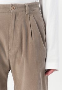 Beige kordfløyelsbukser med plisserte front, klassisk knappelukking og sidelommer, parret med en hvit langermet skjorte.