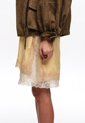 Ręka z czarnym pierścieniem z czerwonym sercem, ubrana w beżową spódnicę z koronkową lamówką oraz brązową zamszową kurtkę z zamkiem błyskawicznym i elastycznym mankietem.