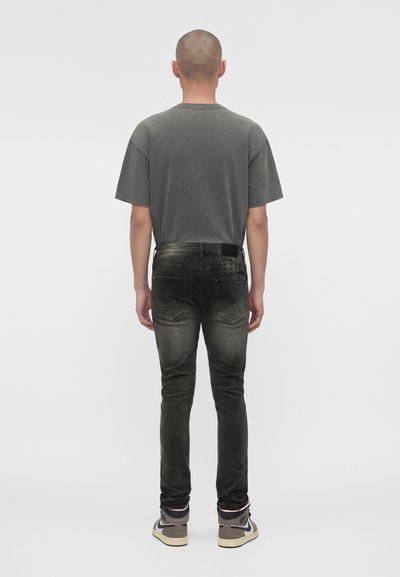 Άνδρας που στέκεται με την πλάτη προς τα έξω φορώντας ξεθωριασμένο μαύρο τζιν, γκρι μπλουζάκι και γκρι ψηλά αθλητικά παπούτσια σε απλό λευκό φόντο.