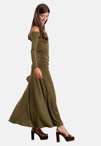 Lungo vestito verde oliva con design off-the-shoulder, dettagli arricciati e gonna fluente. Abbinato a sandali con tacco blocco marrone scuro.