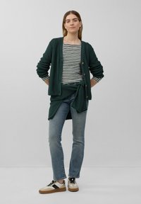 Dunkelgrüne Strickjacke mit Knopfdetails über einem gestreiften Hemd. Dazu werden blaue Jeans und beigefarbene Sneakers kombiniert. Ein schlichter Hintergrund.