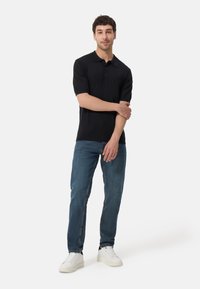 Svart kortärmad polo med krage, tillsammans med blå denimjeans. Modellen bär vita sneakers. Enkel, figurnära design.