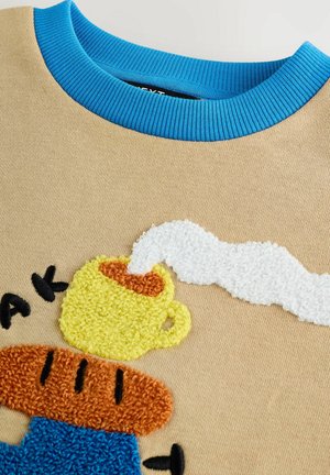Sweatshirt beige avec un col côtelé bleu, présentant une broderie texturée et colorée d'une tasse jaune et d'une tartine brune avec de la vapeur.